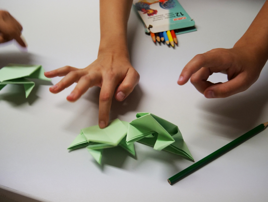KI generiert: Auf dem Bild falten zwei Personen Origami-Frösche aus grünem Papier. Im Hintergrund sind Buntstifte und ein Malbuch zu sehen.