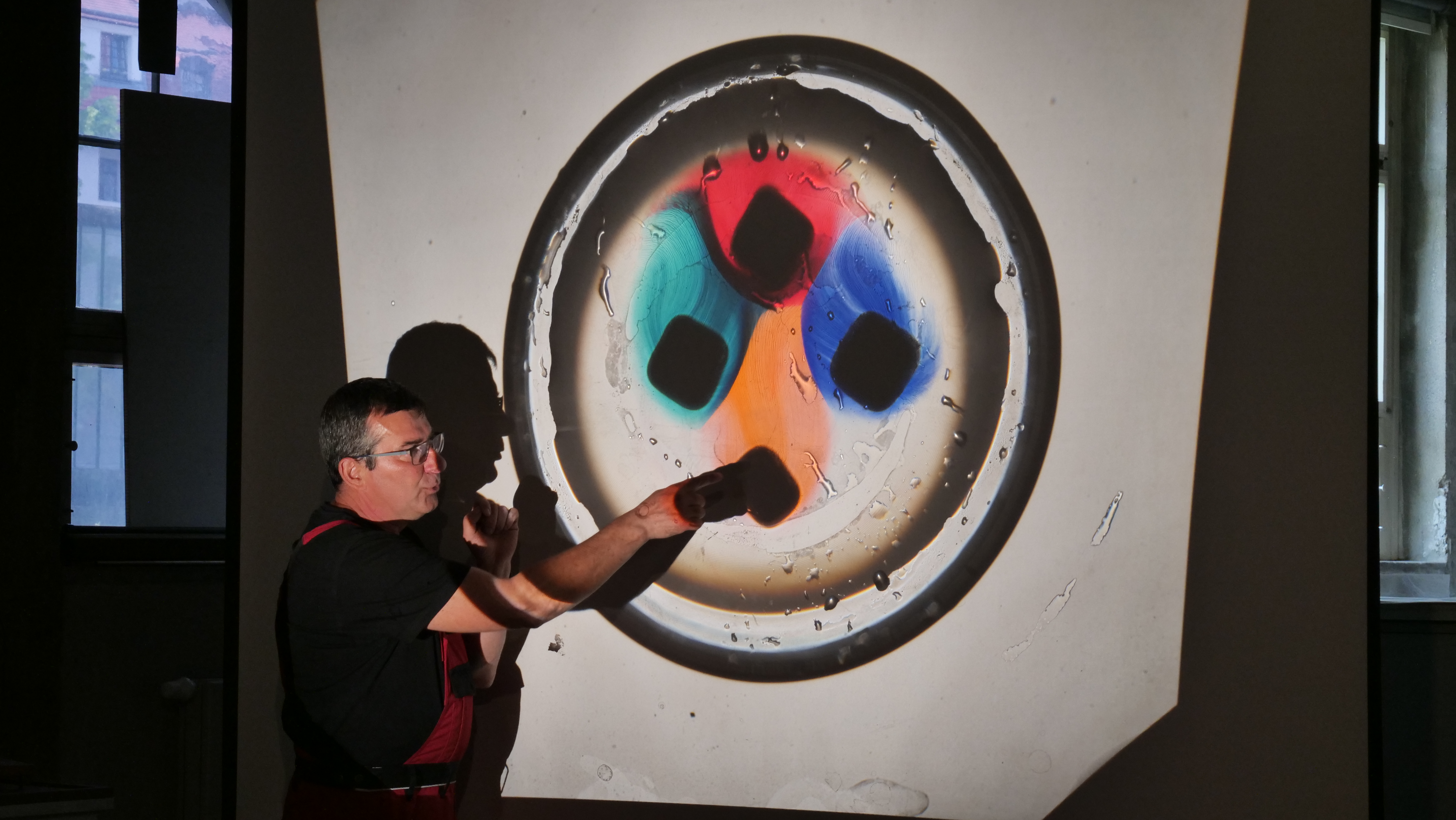 KI generiert: Ein Mann steht vor einer Projektion, die ein kreisförmiges Objekt mit bunten Segmenten zeigt, und zeigt darauf. Die Szene wirkt wie eine Präsentation oder ein Experiment in einem dunklen Raum.