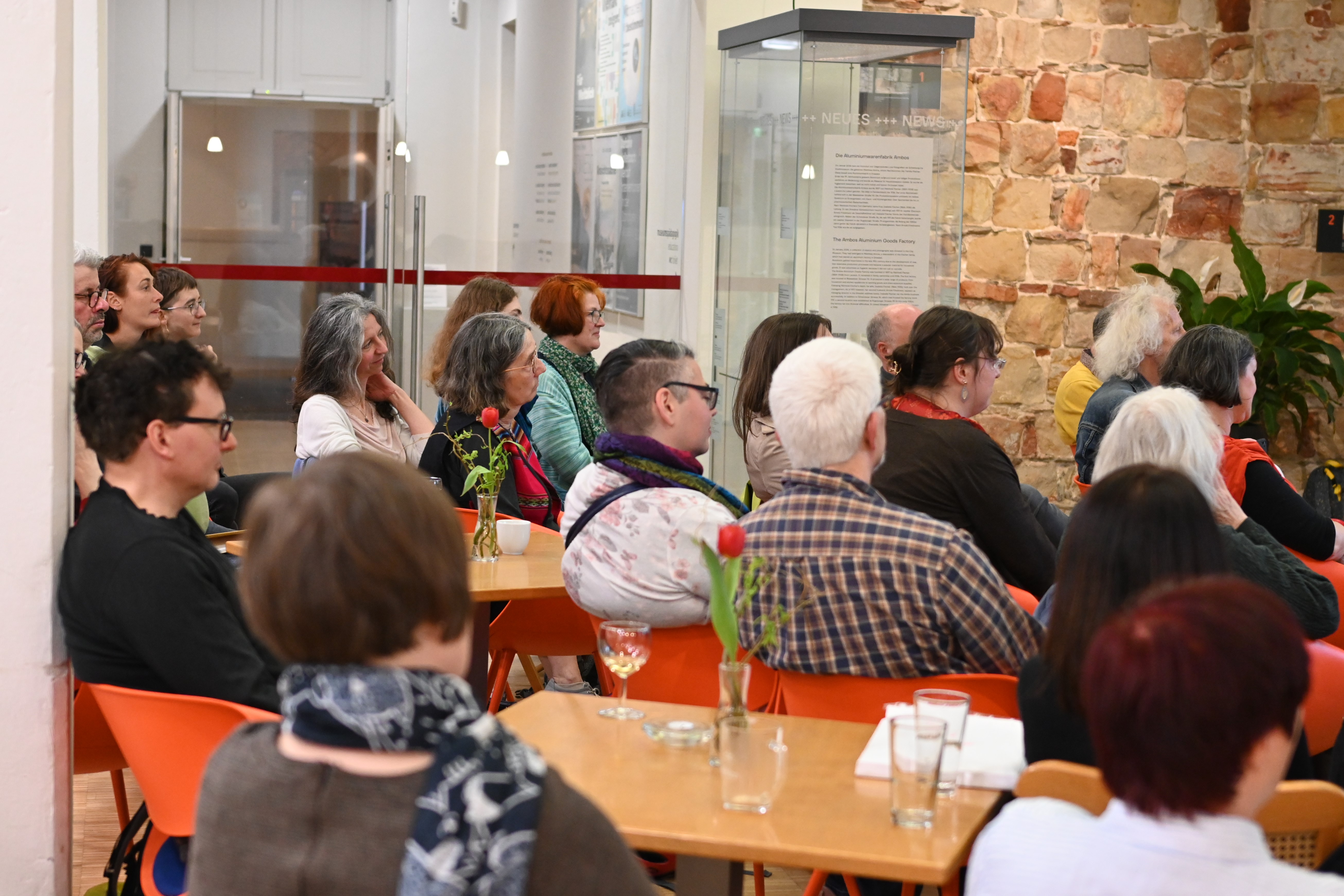 KI generiert: Das Bild zeigt eine Gruppe von Menschen, die aufmerksam einer Präsentation oder Rede innerhalb eines Raumes mit heller Beleuchtung und sichtbarer Steinwand folgen. Auf den Tischen stehen Getränke und Blumen, was auf eine informelle oder gesellige Veranstaltung hindeutet.