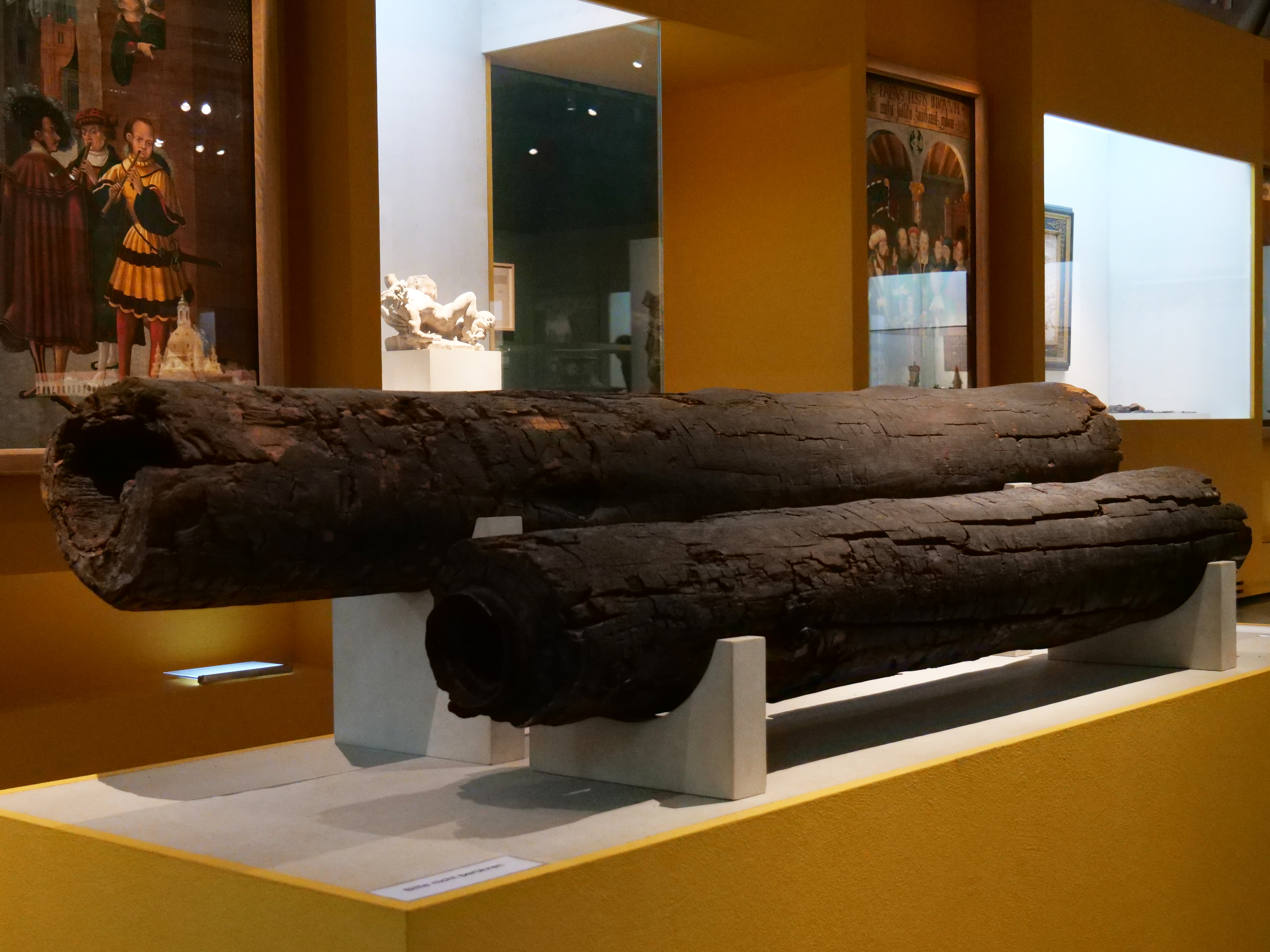 AI generated: The image shows two old wooden water pipes displayed on a pedestal in a museum. They are clearly aged and have a dark, weathered surface.