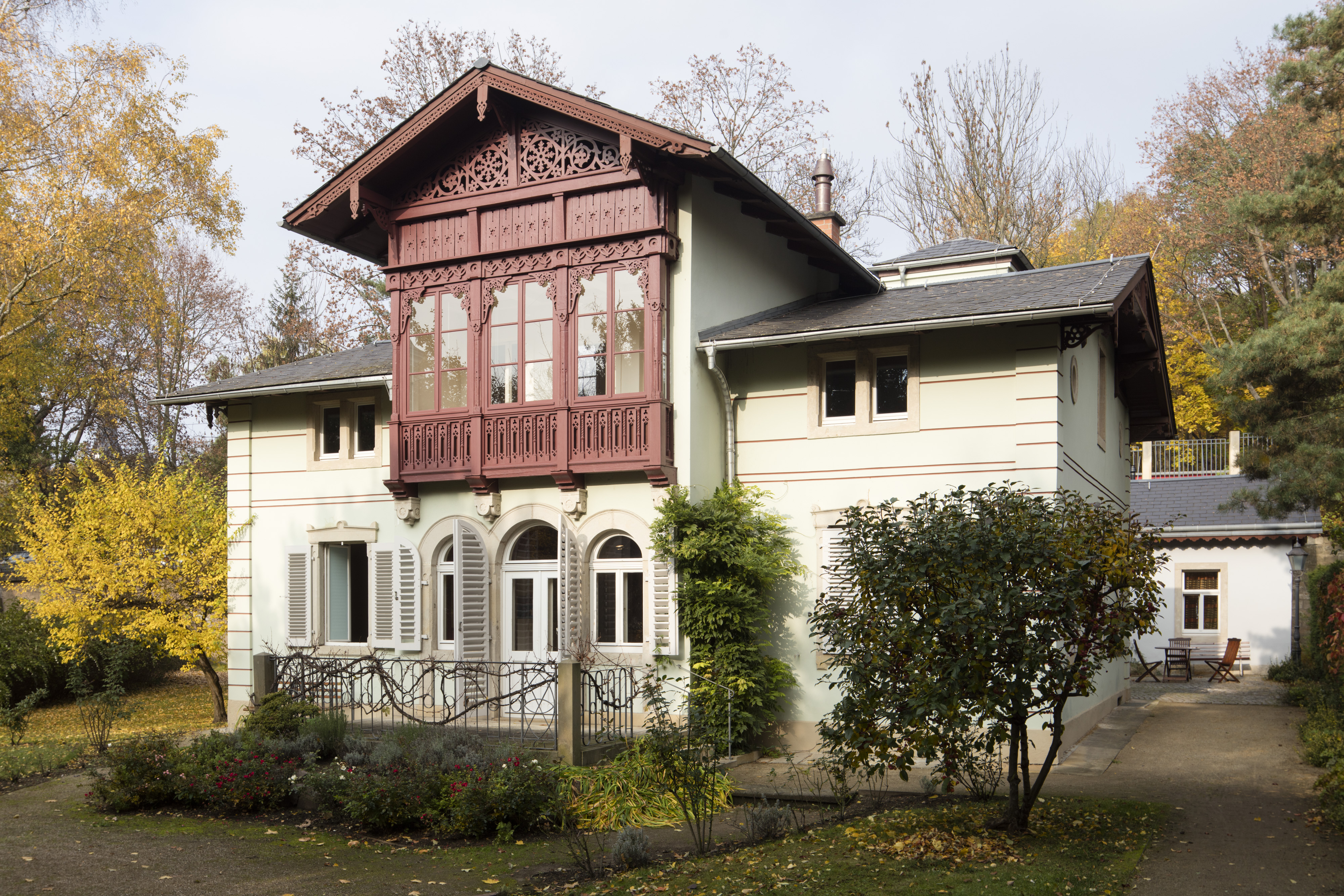 KI generiert: Das Bild zeigt ein charmantes, zweistöckiges Gebäude mit einer detaillierten, roten Holzbalkon-Überdachung und einem gepflegten Garten im Vordergrund. Bäume mit Herbstlaub umgeben das Haus und tragen zur idyllischen Atmosphäre bei.