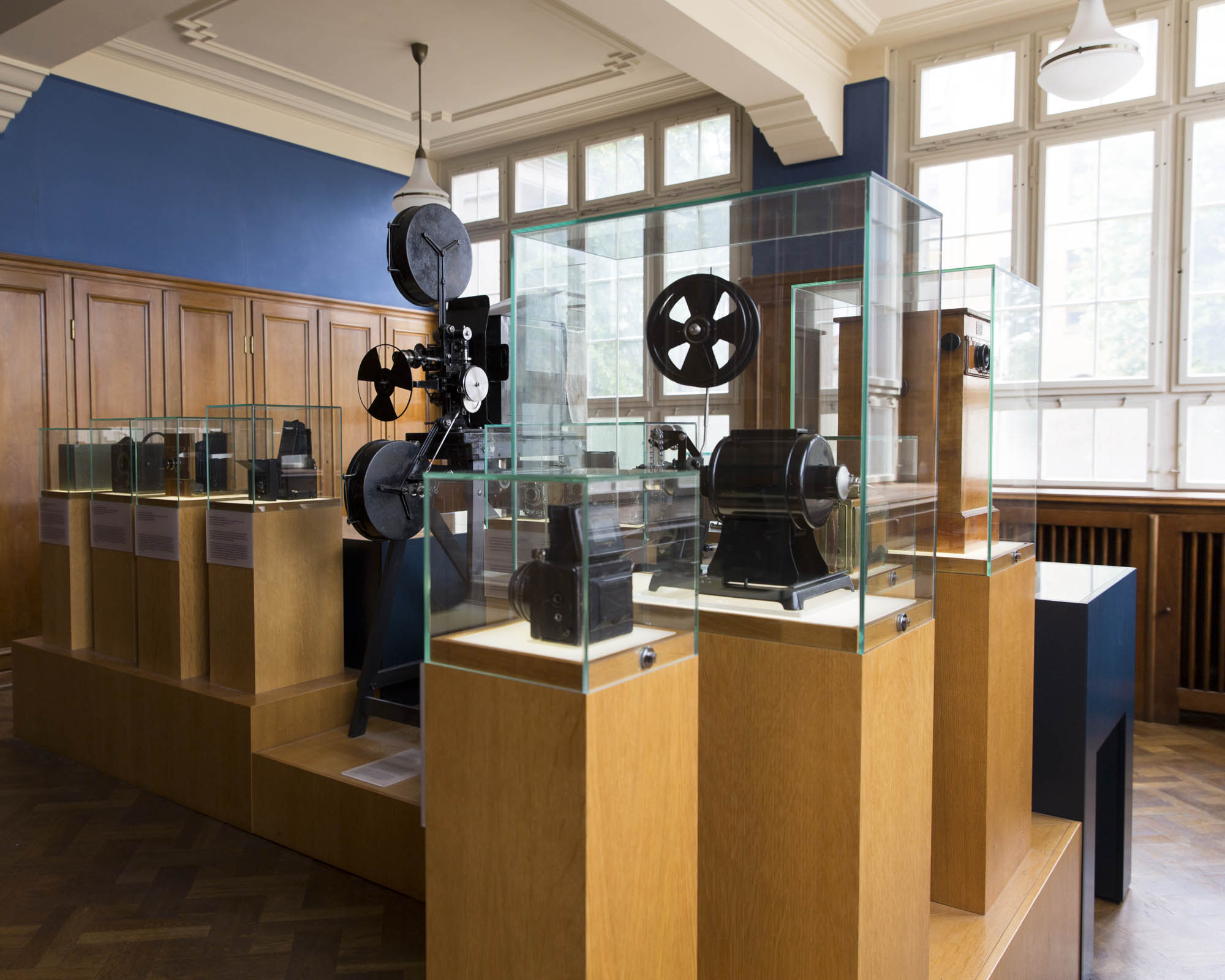 KI generiert: Das Bild zeigt eine Ausstellung historischer Filmprojektoren und Kameras in einem Museum. Die Geräte sind in Holzvitrinen mit Glasabdeckungen ausgestellt.