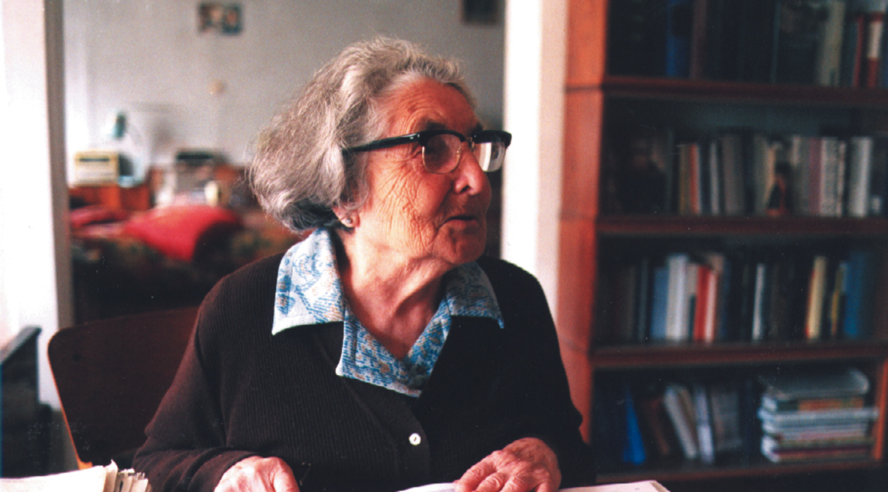 KI generiert: Das Bild zeigt eine ältere Frau, die an einem Tisch sitzt und in einem Buch blättert. Im Hintergrund ist ein Bücherregal zu sehen.