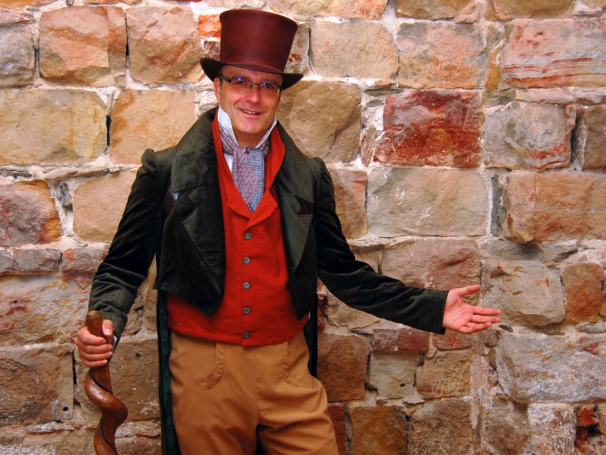 KI generiert: Ein Mann steht lächelnd in historischer Kleidung mit Zylinderhut und Gehstock vor einer Steinmauer. Er trägt einen roten Weste und braune Hosen.