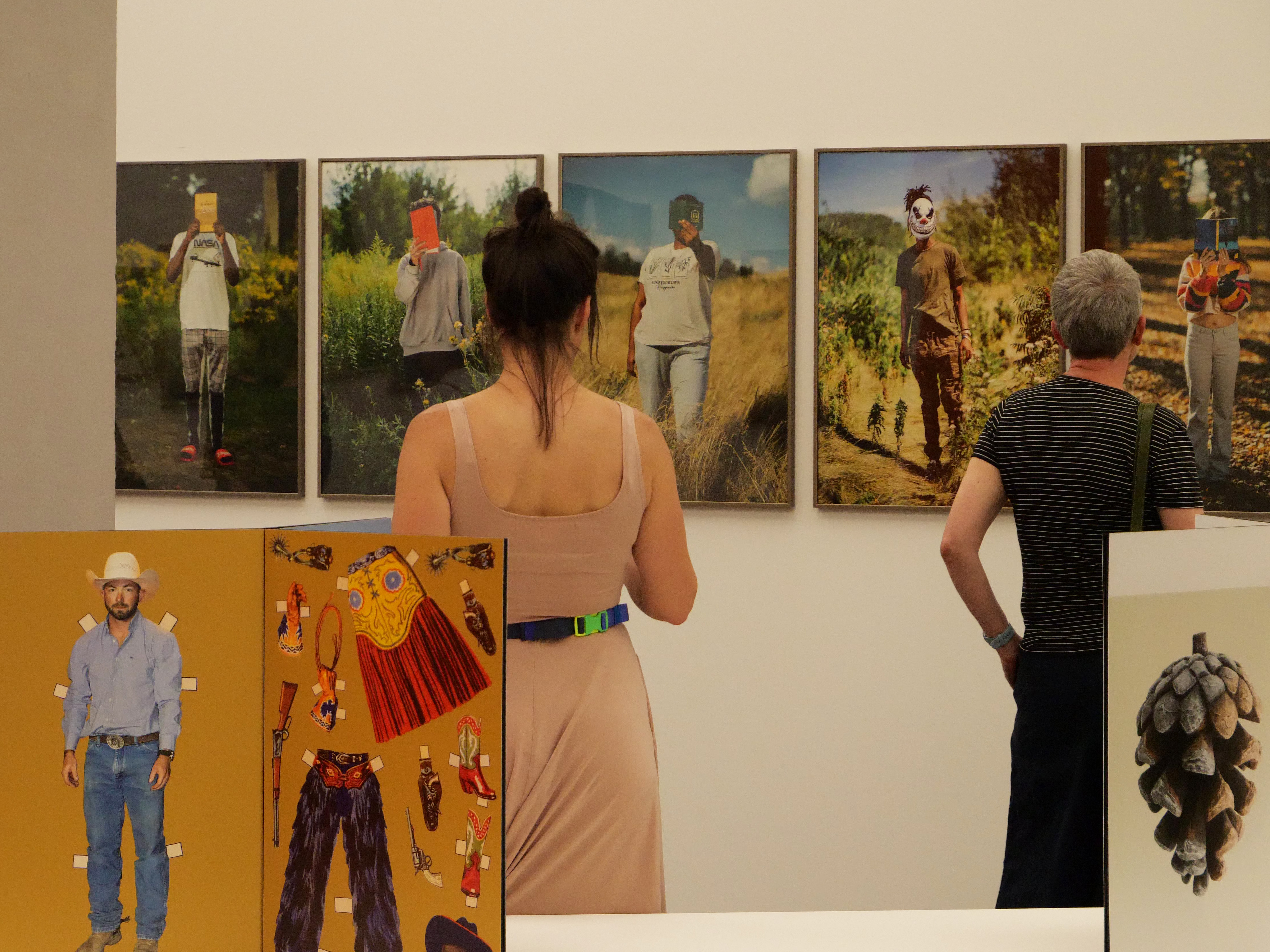 KI generiert: Das Bild zeigt zwei Personen, die sich in einer Kunstgalerie mehrere Fotografien an der Wand ansehen. Die ausgestellten Bilder zeigen Menschen mit maskierten Gesichtern in verschiedenen Umgebungen.