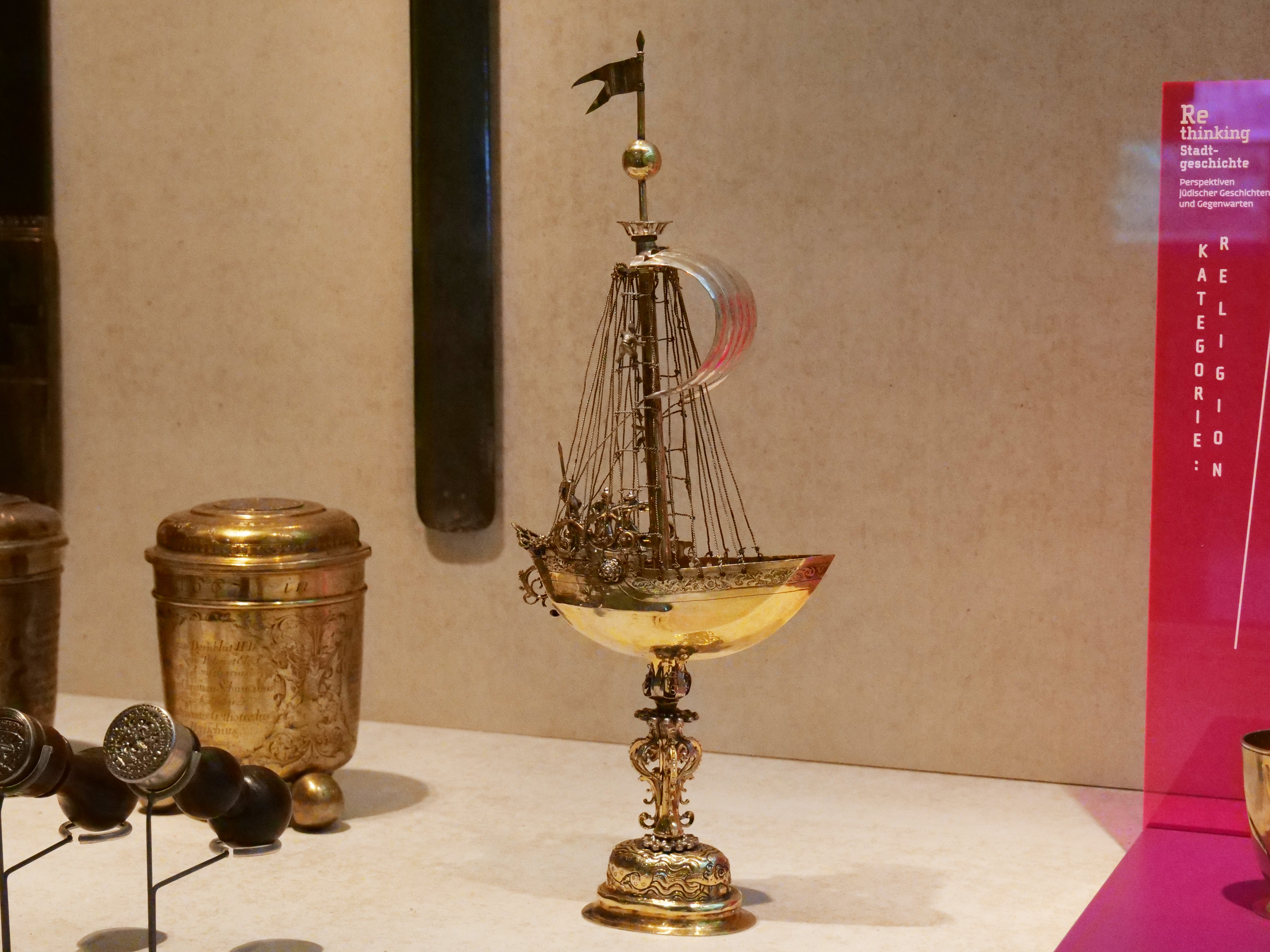 KI generiert: Das Bild zeigt ein kunstvoll verziertes Trinkgefäß in Form eines Schiffs auf einem Sockel, ausgestellt in einem Museum. Um das Schiff herum sind andere antike Gegenstände zu sehen.