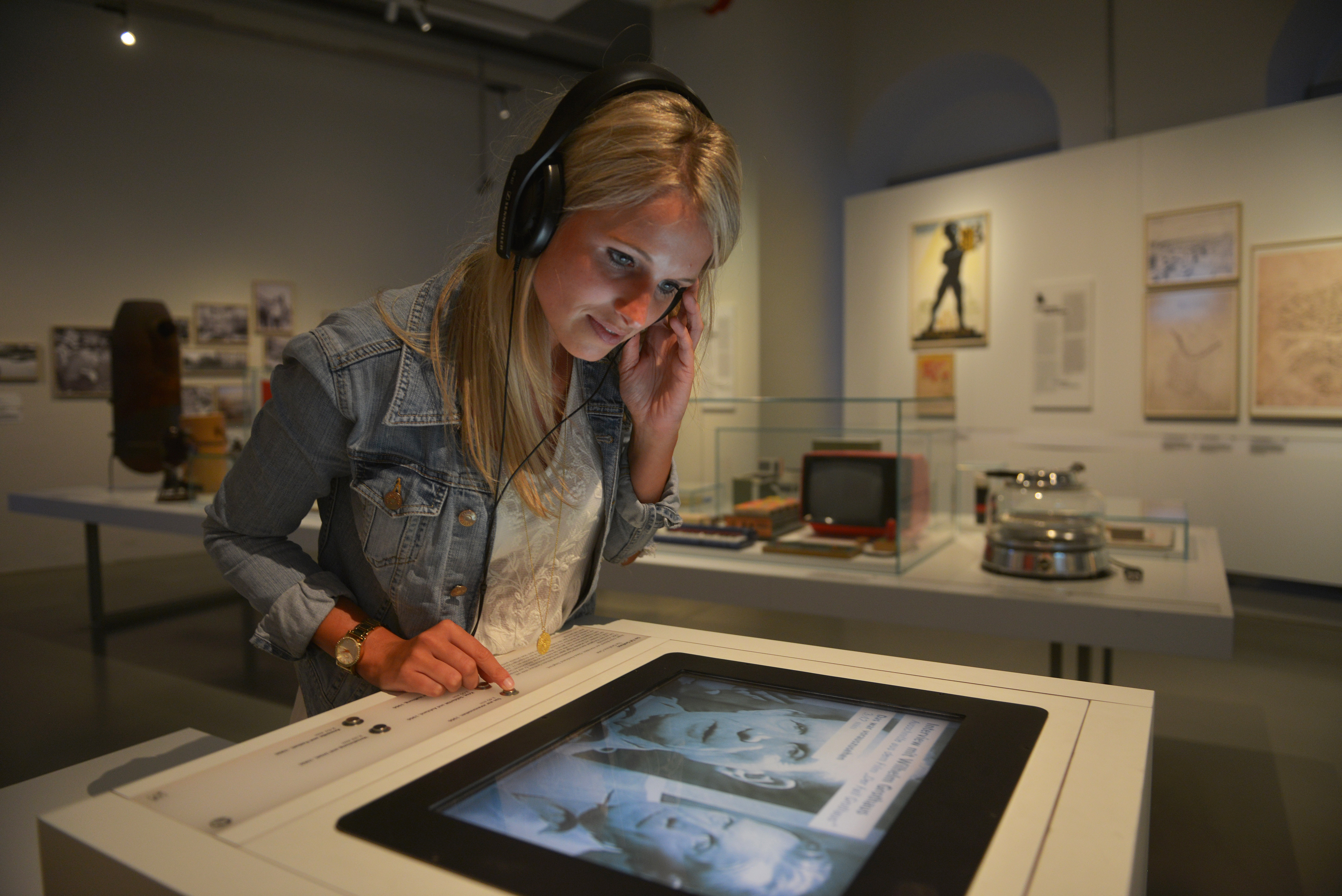AI generated: The image shows a woman wearing headphones and looking at a screen in a museum. The background shows various exhibits and photos.