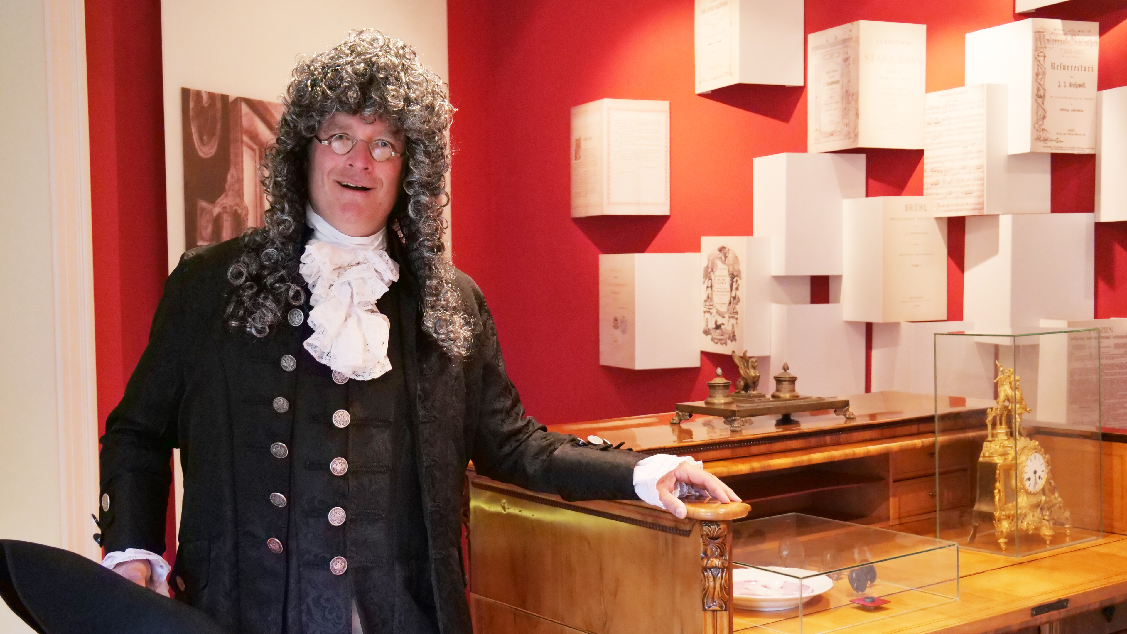 KI generiert: Ein Mann in historischer Kleidung steht in einem Museum vor einer Ausstellung mit roten Wänden und antiken Gegenständen in Vitrinen. Die Ausstellung beinhaltet alte Dokumente und ein aufwendig gestaltetes Uhrwerk.