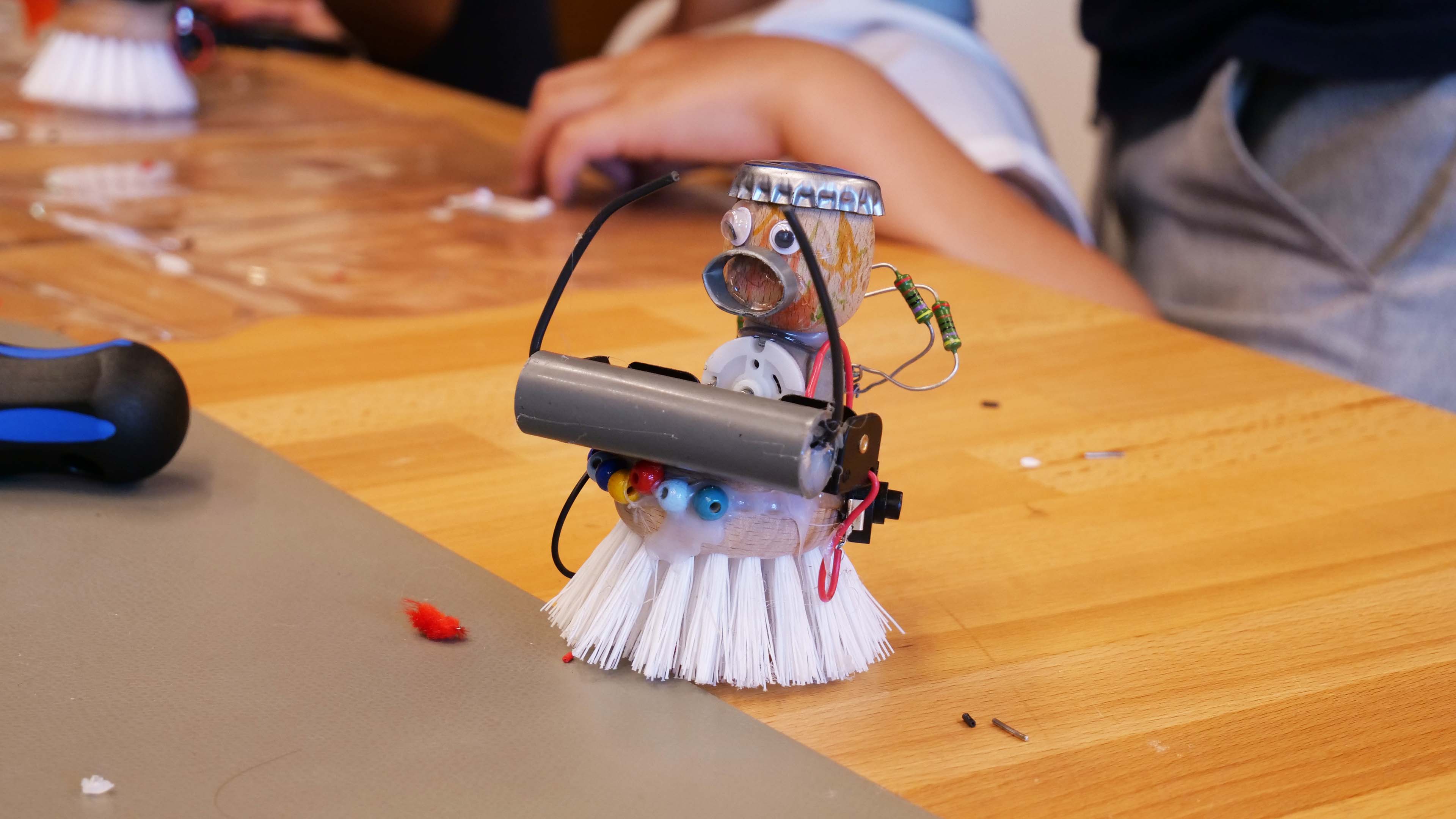 KI generiert: Das Bild zeigt einen kleinen, selbstgebauten Roboter, der aus einem Handfeger, einem Motor und verschiedenen Elektronikkomponenten besteht. Im Hintergrund sind Hände und Teile weiterer ähnlicher Projekte zu sehen.
