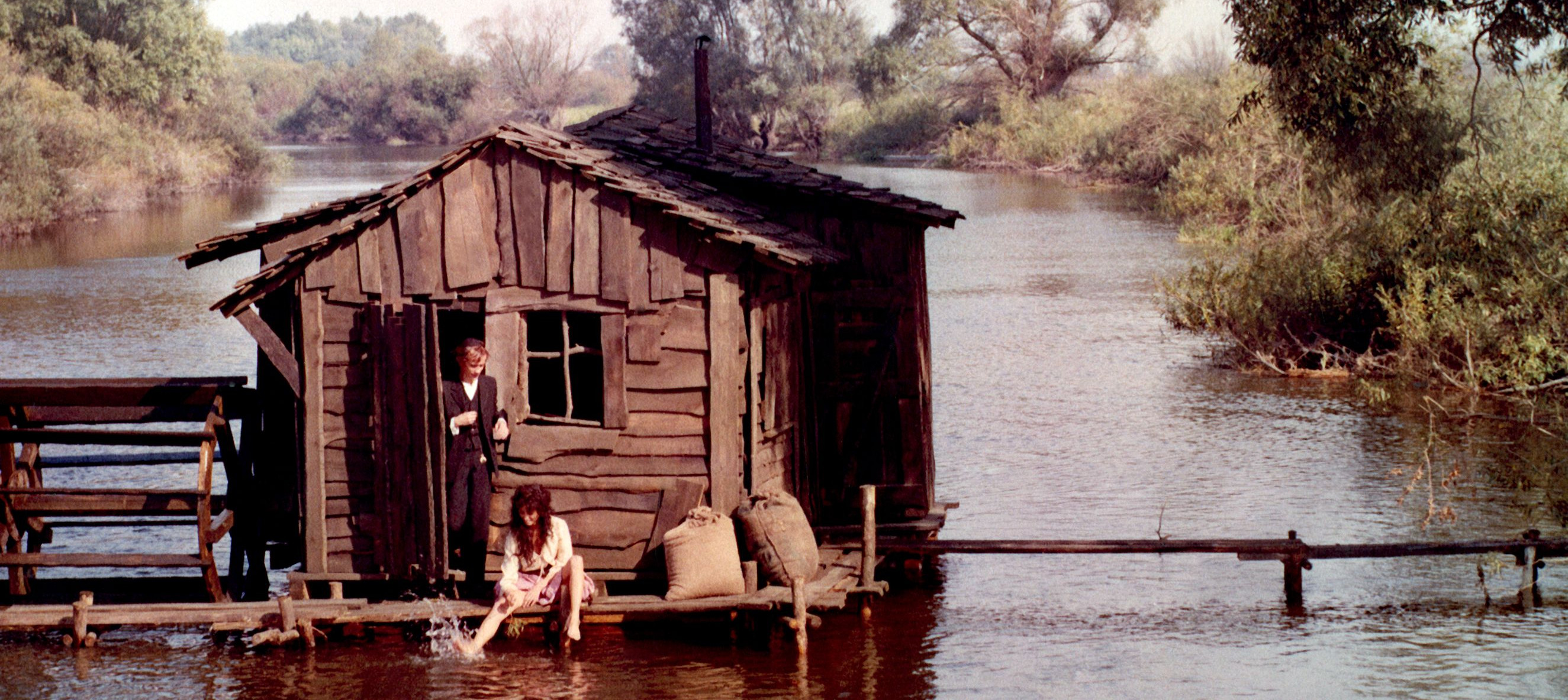 KI generiert: Das Bild zeigt eine hölzerne Hütte, die auf einem See oder Fluss steht, mit zwei Personen: Eine sitzt am Rand und planscht mit den Füßen im Wasser, während die andere in der Tür steht. Im Hintergrund ist üppige Vegetation sichtbar, was eine ruhige, naturverbundene Szene schafft.