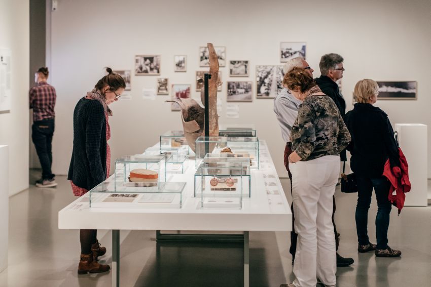 KI generiert: Das Bild zeigt mehrere Menschen, die sich in einem Museum oder einer Ausstellung befinden und die Exponate in gläsernen Vitrinen betrachten. Im Hintergrund sind mehrere Fotografien an der Wand zu sehen.