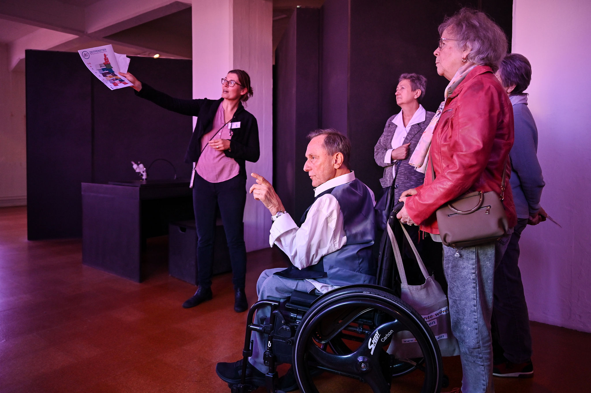 KI generiert: Eine Frau hält ein Blatt Papier hoch und erklärt einer Gruppe von Menschen im Innenraum etwas, darunter ein Mann im Rollstuhl. Die Gruppe ist in ein Gespräch vertieft oder hört aufmerksam zu.