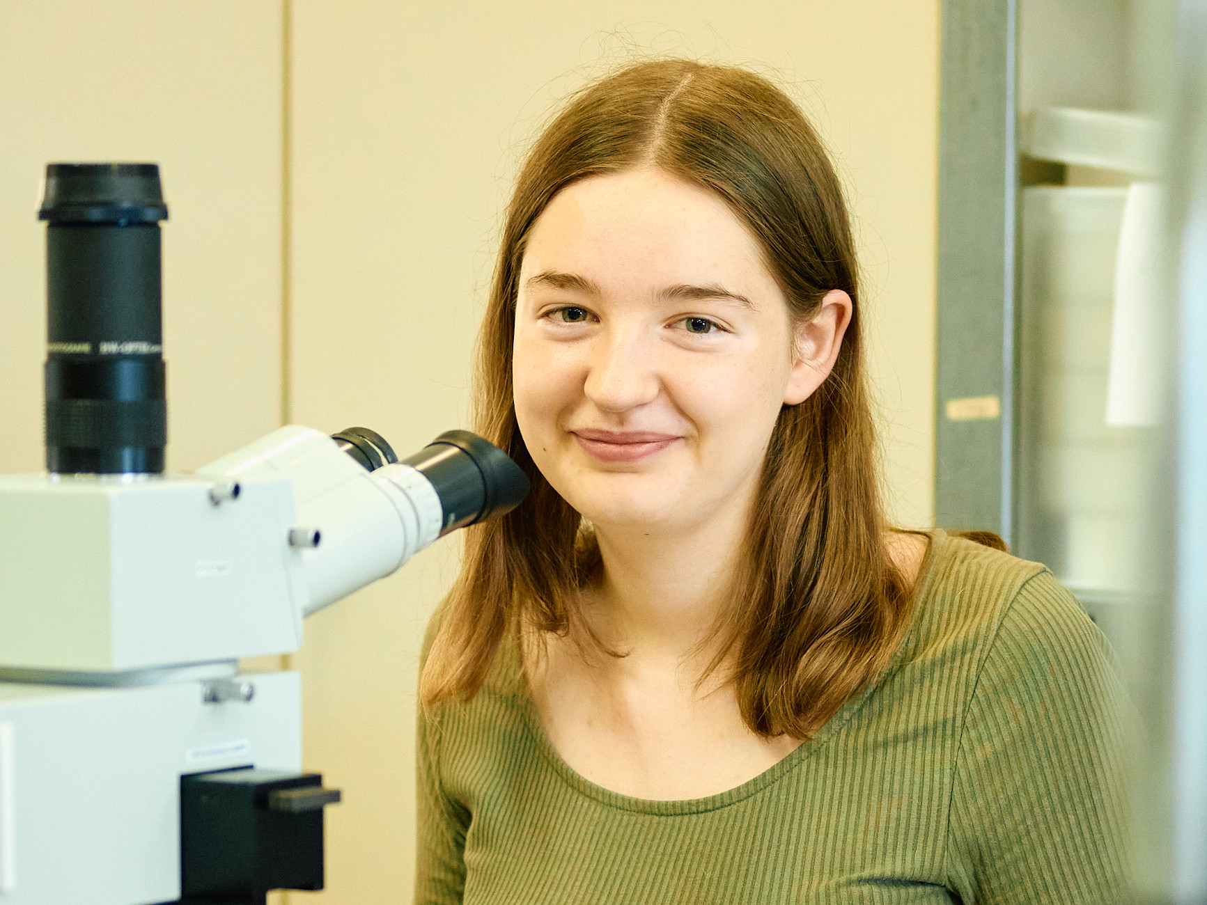 KI generiert: Das Bild zeigt eine junge Frau, die neben einem Mikroskop sitzt und in die Kamera lächelt. Der Hauptinhalt des Bildes besteht aus der Person und dem Mikroskop, was auf eine wissenschaftliche oder Laborumgebung hinweist.