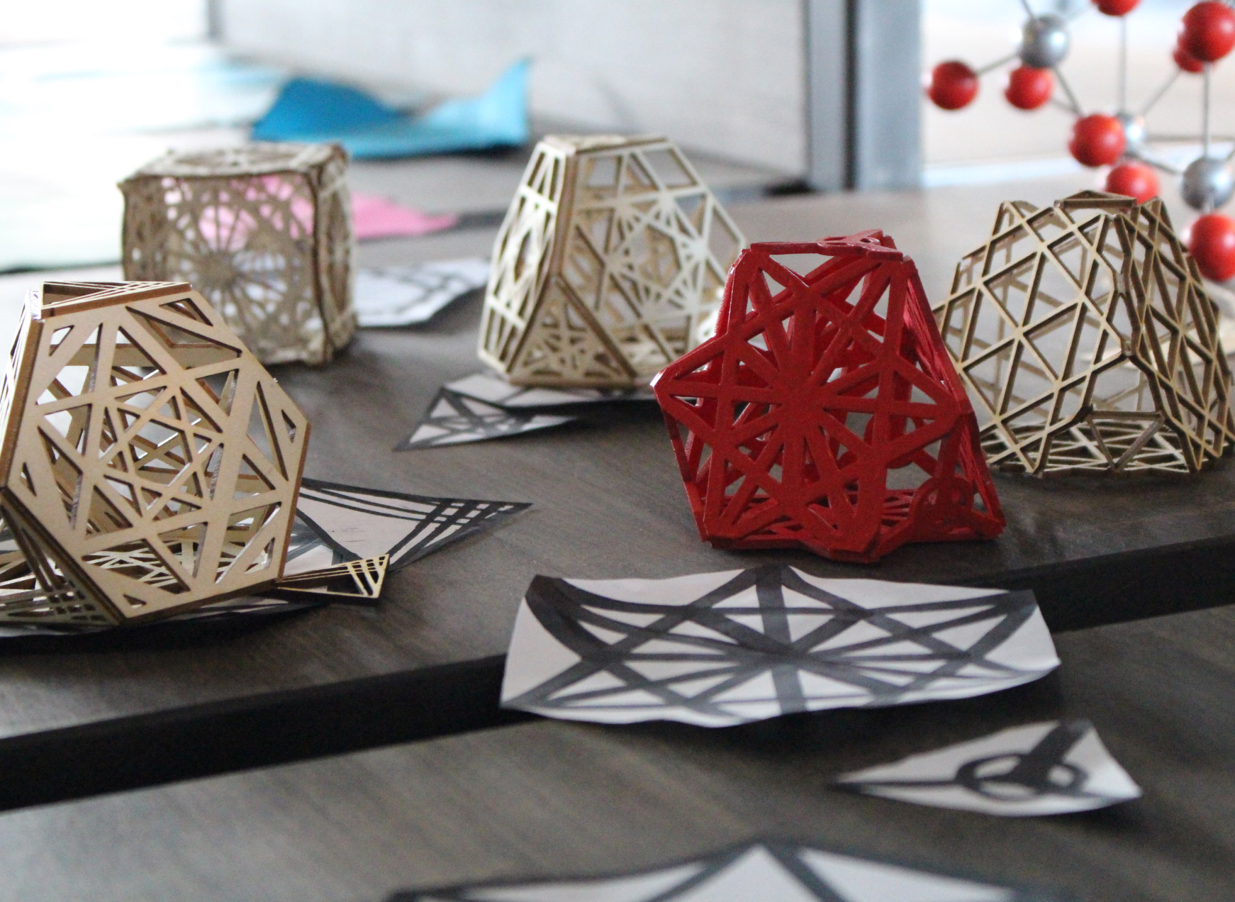 KI generiert: Das Bild zeigt geometrische, gitterförmige Modelle auf einem Tisch, darunter ein rotes und mehrere beige-farbige. Im Hintergrund sind einige Schwarz-Weiß-Zeichnungen von ähnlichen geometrischen Mustern zu sehen.